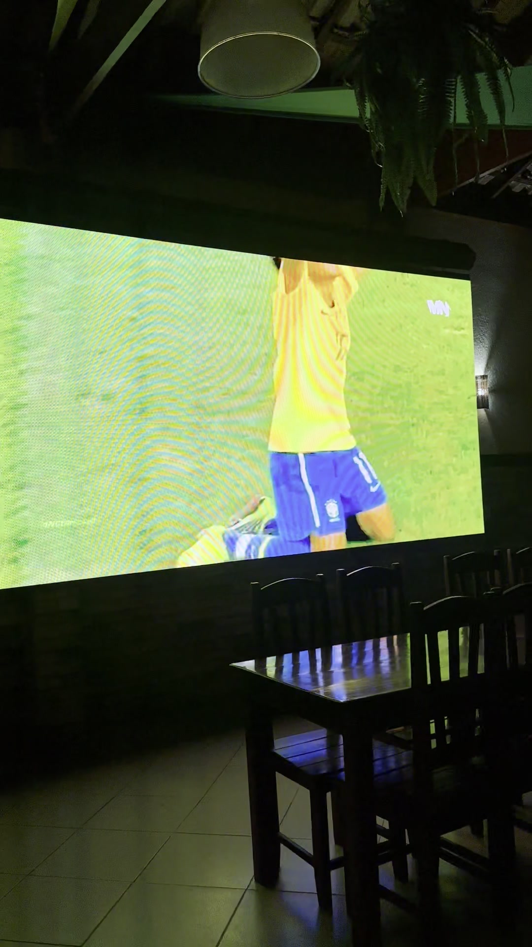 Telão da casa exibindo jogo de futebol em ambiente escuro